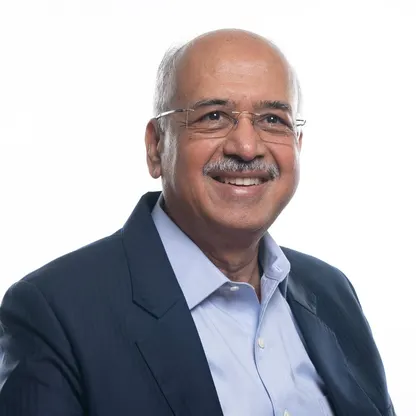 Portrait of Dilip Shanghvi smiling, in a dark jacket and light blue shirt, against a white background.