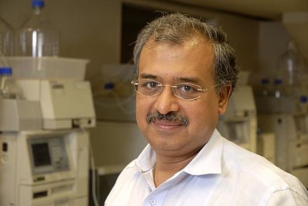 Dilip Shanghvi photographed in a laboratory, wearing a light shirt, with analytical instruments behind him.