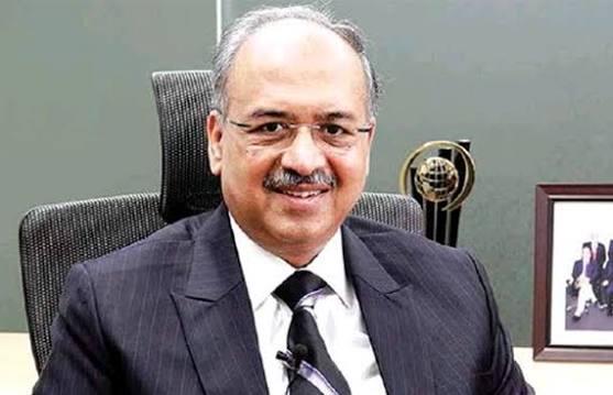 Dilip Shanghvi seated in an office chair in a dark suit, with an award and a framed photograph behind him.