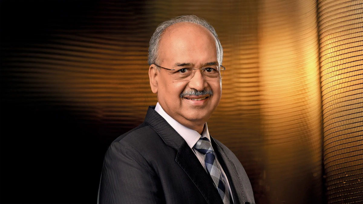 Portrait of Dilip Shanghvi in a dark suit and patterned tie against a warm gold background.