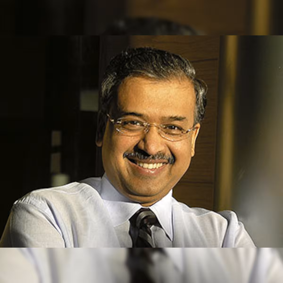 Portrait of Dilip Shanghvi smiling, wearing a light shirt and dark tie, in warm interior light.