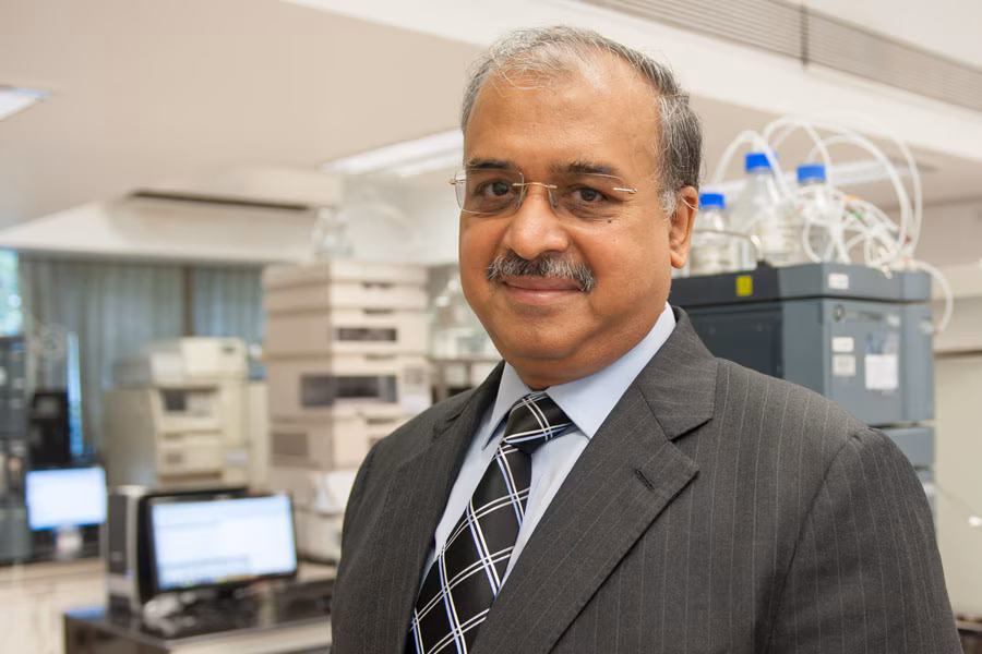 Dilip Shanghvi in a grey suit standing in a laboratory with analytical equipment and computer workstations.